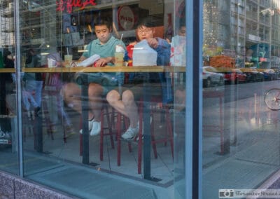 Two young people eating at a counter behind glass, a bicycle and traffic mirrored across it, Fuji X100V