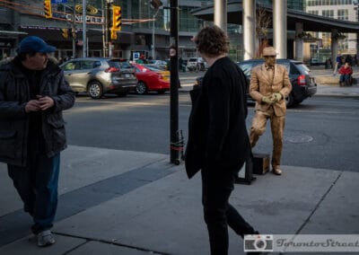 Two people walking past a gold painted performer on the sidewalk, a Cineplex sign behind, Leica Q2