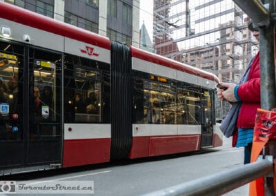 A red and white streetcar at a stop, a woman in a red coat waiting with her phone, Leica Q2