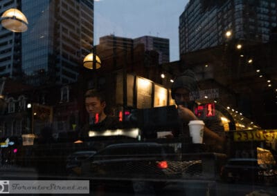 Two people at a coffee counter at dusk, city towers and neon laid over the glass, Leica Q2
