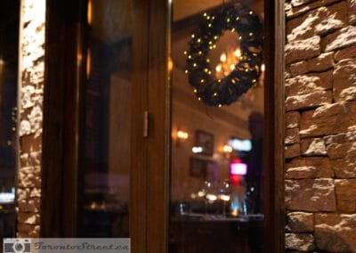 A lit wreath hanging in a doorway, stone wall beside it and a warm dining room beyond, Leica Q2