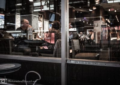 Man on a phone and a woman in leopard print in a bar, a Winners sign backwards on the glass, Leica Q2