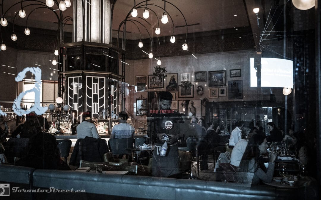 A grand bar at night seen through Toronto windows, deco lamp cluster and a wall of framed pictures, Leica Q2