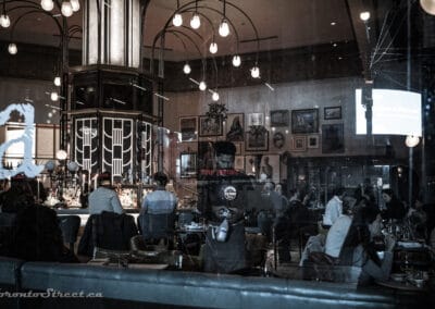 A grand bar at night seen through Toronto windows, deco lamp cluster and a wall of framed pictures, Leica Q2