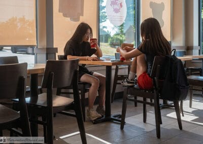 Two teenagers at a sunlit window table with a backpack and drinks, green trees outside, Leica Q3