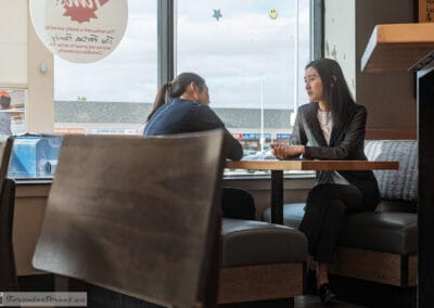 Woman in a black blazer talking with two others in a window booth, a plaza outside, Leica Q3
