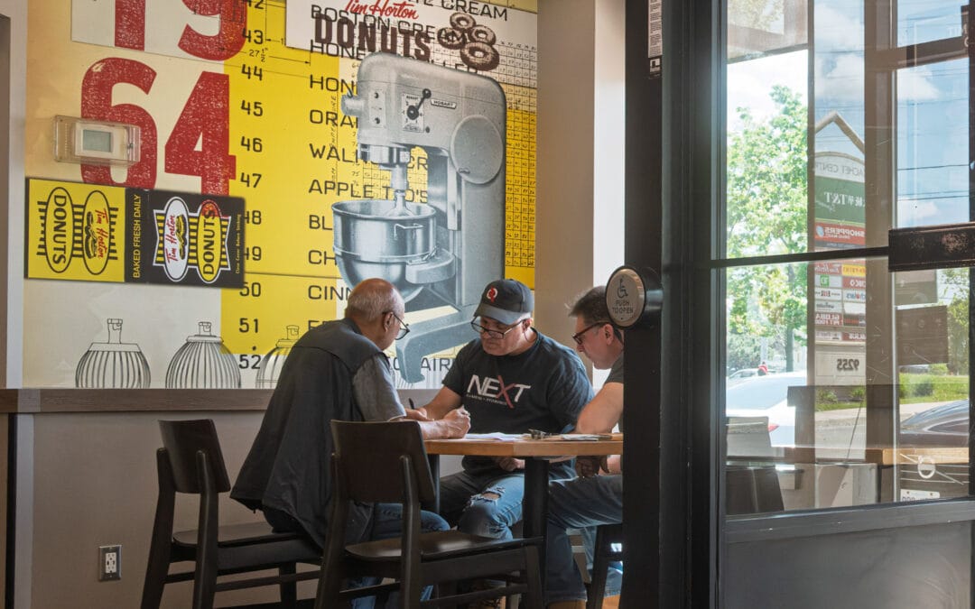 Three men working through paperwork inside Tim Hortons under a yellow 1964 donut price mural, Leica Q3