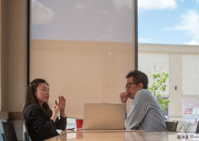 A woman gesturing across a laptop at a man in a checked shirt, sunlit blind behind them, Leica Q3
