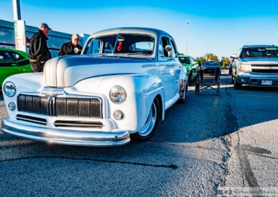 Two men talking at the nose of a white and silver coupe in the Canadian Tire lot, green Challenger behind, Leica Q3