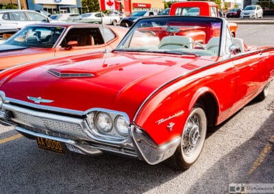 Front three-quarter of the red Bullet Bird on wire wheels and whitewalls, flag flying, orange coupe behind, Leica Q3
