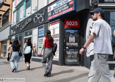 Busy Yonge Street sidewalk outside a walk-in clinic and an optician, people passing both ways, Toronto, Leica Q3