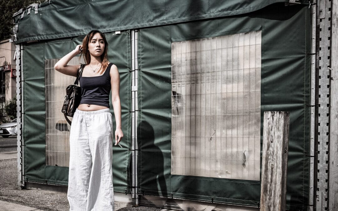 Young woman in a black top and wide white trousers against a green canvas hoarding on a Toronto street, Leica Q3