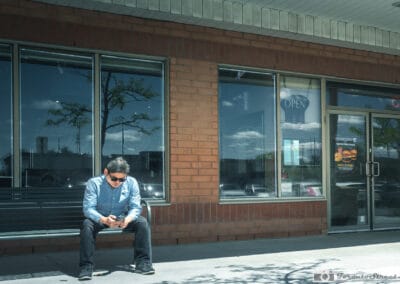 Man in a blue shirt and sunglasses hunched on a bench outside a brick restaurant, thumbs on a small screen, Leica Q3