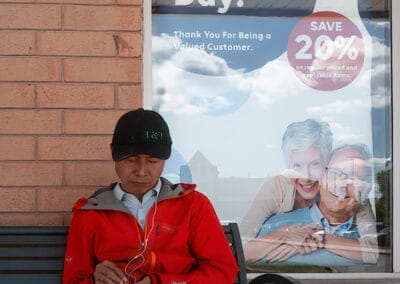 Man in a red jacket untangling white earbuds on a bench under a Seniors' Day poster, GTA plaza, Leica Q3