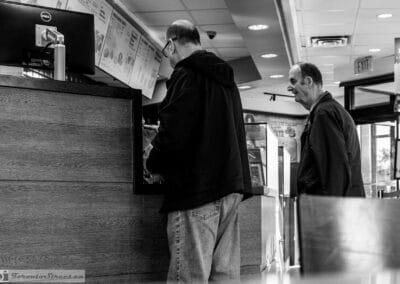 Two older men at a Tim Hortons counter in black and white, one waiting while the other pays, Leica Q3