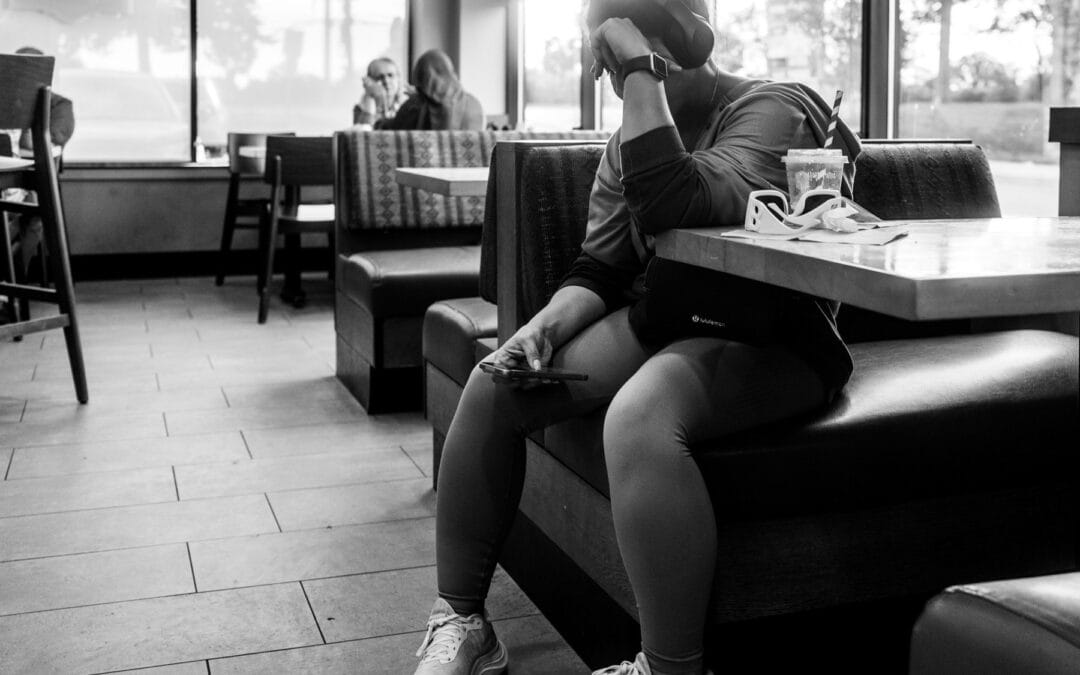 Woman in a coffee shop booth, headphones on, phone held low, a couple on their phones in the booth behind, Toronto, Leica Q3