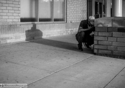 Man crouched at the base of a brick pillar with his phone, hand over his mouth, long evening shadow on the wall, Leica Q3