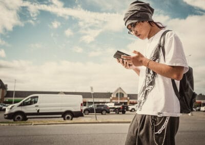Young man in a grey beanie walking across a suburban plaza lot, head down over the screen in both hands, Leica Q3