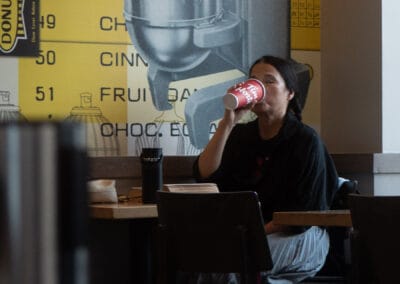 Woman tipping back a Tim Hortons cup under the yellow 1964 price list mural, her own thermos beside her, Leica Q3