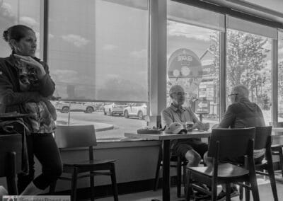 Monochrome frame of two older men at a window table, a woman standing at left with her hand at her chin, Leica Q3