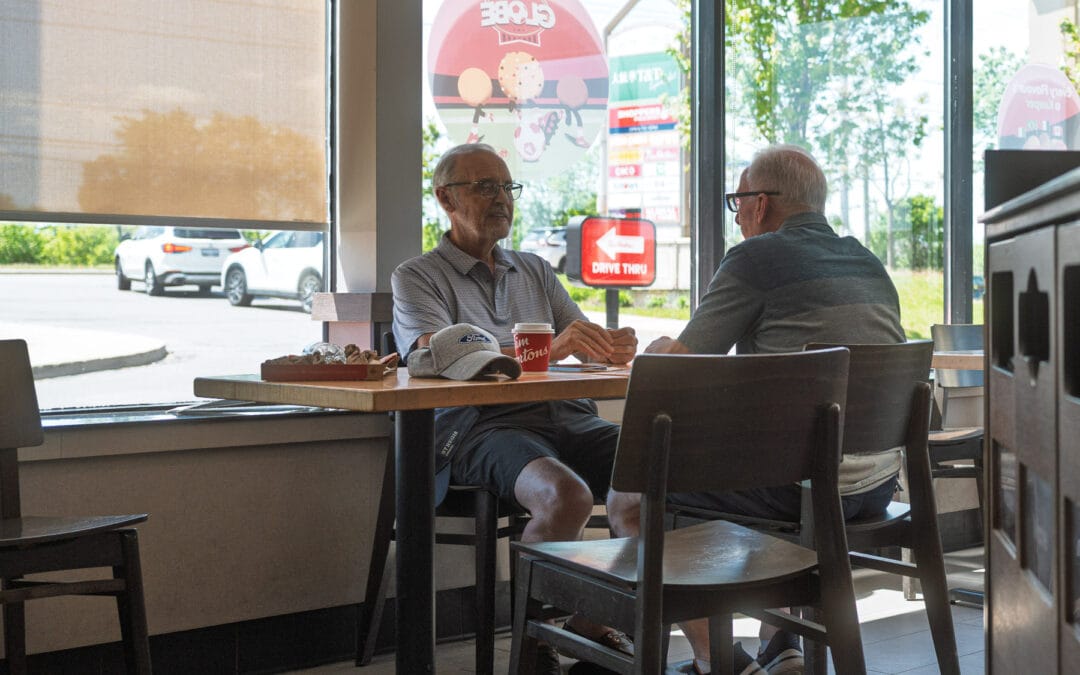 Two older men at a Tim Hortons window table, red drive-thru sign and green trees outside on a Sunday, GTA, Leica Q3