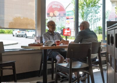 Two older men at a Tim Hortons window table, red drive-thru sign and green trees outside on a Sunday, GTA, Leica Q3