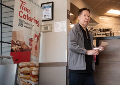 Older man in a grey jacket with a coffee and paper bag looks straight at the lens beside a catering banner, Leica Q3