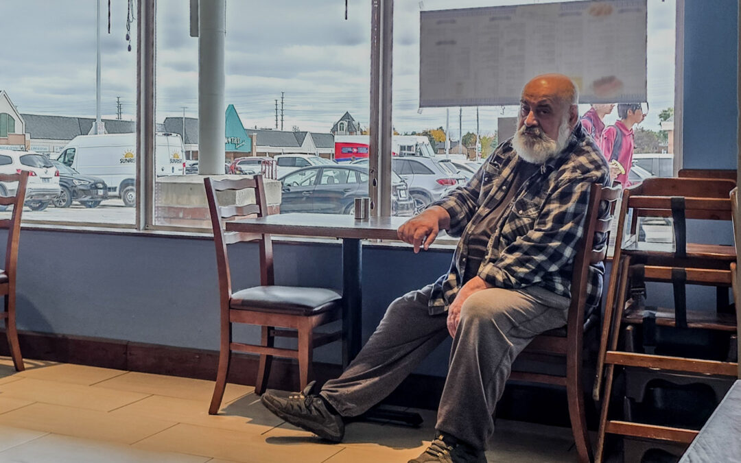 Bearded man in a plaid shirt sitting sideways by a restaurant window, looking at the camera, Toronto, taken on a phone