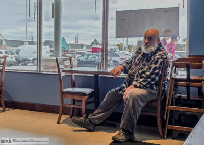 Bearded man in a plaid shirt sitting sideways by a restaurant window, looking at the camera, Toronto, taken on a phone