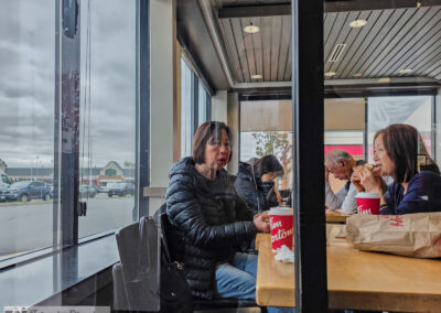 Two women talking over red coffee cups at a window table, seen through glass, plaza lot outside, Toronto, taken on a phone